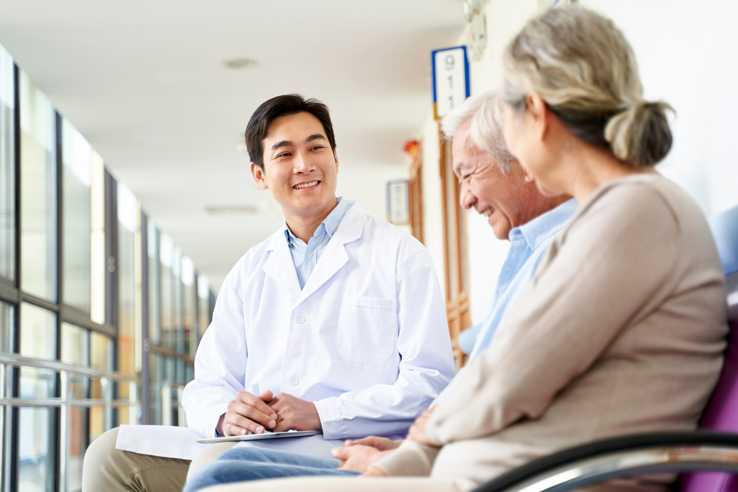 friendly young asian doctor talking to old couple in hospital hallway